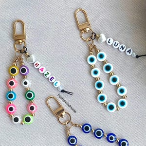 Op de afbeelding: Drie goudkleurige sleutelhangers met karabijnsluiting en lussen van kleurrijke boze oog kralen. Eén sleutelhanger heeft witte letterkralen die HAZEL spellen, een andere LUNA, en de derde heeft blauwe boze oog kralen met gouden tussenstukjes.