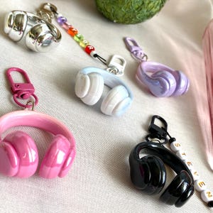 Puede incluir: Una colecci&oacute;n de llaveros de auriculares coloridos en plata, rosa, azul, morado y negro. Cada llavero tiene un cierre de mosquet&oacute;n y algunos tienen cuentas con letras. Los auriculares son brillantes y detallados.