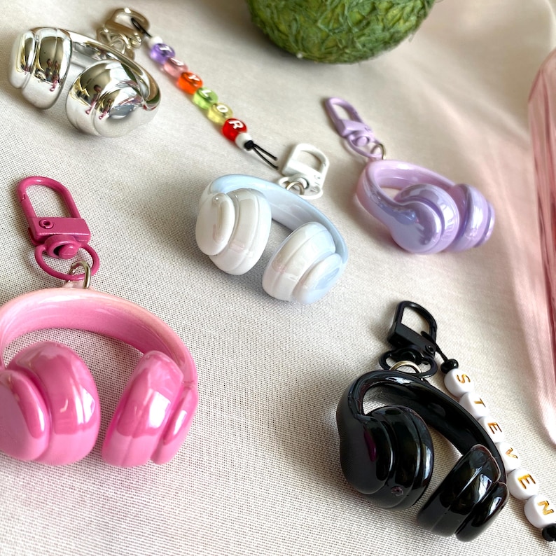 Puede incluir: Una colecci&oacute;n de llaveros de auriculares coloridos en plata, rosa, azul, morado y negro. Cada llavero tiene un cierre de mosquet&oacute;n y algunos tienen cuentas con letras. Los auriculares son brillantes y detallados.