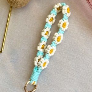 May include: A white and blue macrame keychain with a gold key ring. The keychain is made of a series of daisy-shaped knots with yellow centers.