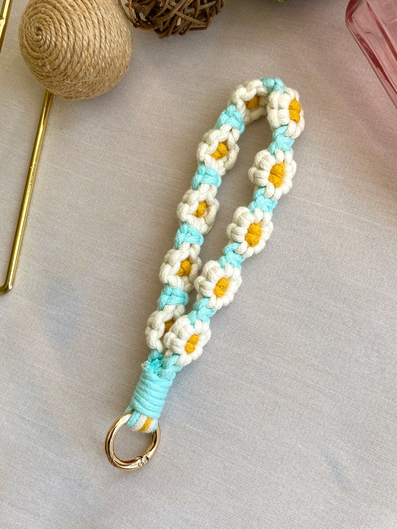May include: A white and blue macrame keychain with a gold key ring. The keychain is made of a series of daisy-shaped knots with yellow centers.