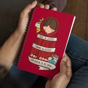 Puede incluir: Cuaderno espiral rojo con una ilustración de dibujos animados de una niña con una capa roja con capucha. El texto en el cuaderno dice "Come un poco, bebe un poco y escucha mi pena."