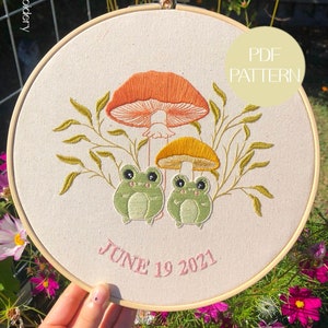 May include: Embroidered hoop art featuring two green frogs sitting under a large orange mushroom and a smaller yellow mushroom. The hoop is surrounded by green leaves and the text "JUNE 19 2021" is embroidered in pink below the frogs. A white label with the text "PDF PATTERN" is in the upper right corner of the hoop.