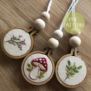 May include: Three small, round embroidery hoops with botanical designs. One features a red mushroom with white spots, another a sprig of purple flowers, and the third a green leafy branch. Each is attached to a string of wooden beads. The text "PDF PATTERN" is visible.