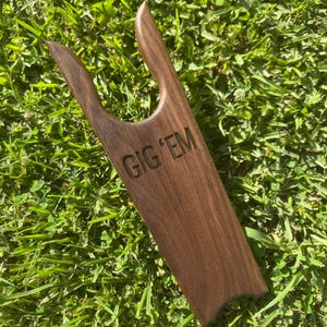 May include: A wooden boot jack with the text "GIG 'EM" carved into the wood. The boot jack is brown and sits on a bed of green grass. The words "CUSTOM BOOT JACKS" and "PERSONALIZED TO ORDER" are in the image.
