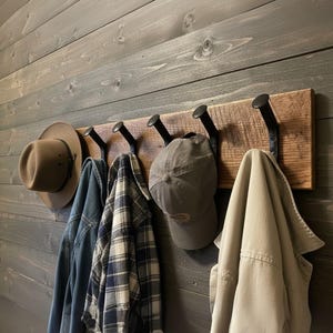 May include: A rustic wooden coat rack with five black metal hooks. Various items hang from the hooks, including a brown hat, a denim jacket, a plaid shirt, a gray baseball cap, and a tan jacket. The rack is mounted on a gray wooden wall.