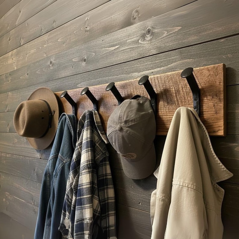 May include: A rustic wooden coat rack with five black metal hooks. Various items hang from the hooks, including a brown hat, a denim jacket, a plaid shirt, a gray baseball cap, and a tan jacket. The rack is mounted on a gray wooden wall.