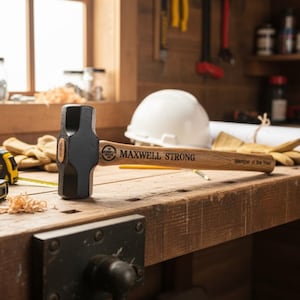 May include: A black sledgehammer with a wooden handle, engraved with "MAXWELL STRONG" and "Member of the Year." The hammer rests on a wooden workbench, alongside a white hard hat, gloves, and a measuring tape.