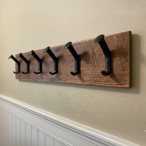May include: A rustic wooden coat rack with six black metal hooks. The wood has a natural, weathered appearance with visible grain and knots. The hooks are evenly spaced and curved, providing a functional and decorative storage solution for a hallway or entryway.