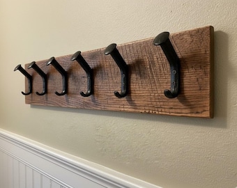 May include: A rustic wooden coat rack with six black metal hooks. The wood has a natural, weathered appearance, and the hooks are evenly spaced along the length of the board. The rack is mounted on a beige wall.