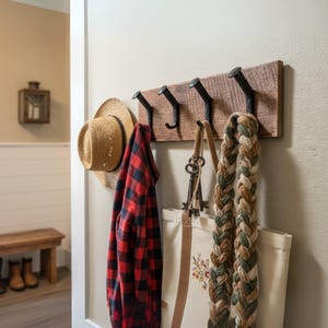 May include: A rustic wooden wall-mounted coat rack with four black metal hooks. A straw hat, a red and black plaid shirt, a canvas tote bag, and a braided scarf hang from the hooks. The rack is mounted on a light beige wall.