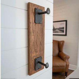 May include: A rustic wall-mounted coat rack made of reclaimed wood. The brown wooden plank features two dark metal hooks attached with square plates. The rack is mounted on a white wall, showcasing a blend of industrial and farmhouse design.
