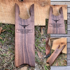 May include: A wooden boot jack with a cutout for the heel of a boot. The boot jack is engraved with the text "Magnolia Oaks Farm & Ranch" and "Houston, Texas".