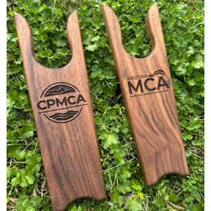 May include: Two wooden boot jacks with a dark brown finish. One has the text "CPMCA" and the other has "ARIZONA MCA Mechanical Contractors Association" engraved on them. The boot jacks are on a bed of green grass.