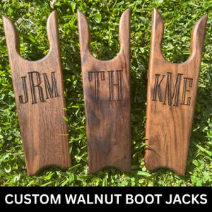 May include: Three custom walnut boot jacks with engraved initials: JRM, TH, and KME. The wooden boot jacks are displayed on a bed of green grass. The text "CUSTOM WALNUT BOOT JACKS" is at the bottom of the image.