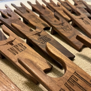 May include: A row of wooden gun racks with personalized names, including "WEB", "SGS", "OCP", and "AD". Each rack features a carved Texas longhorn design.