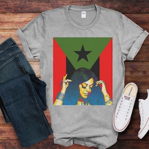 Puede incluir: Una camiseta gris con un gráfico de una mujer con cabello negro largo que lleva una camisa estampada en rojo, verde y negro. El gráfico incluye una estrella y la mujer se está sosteniendo las manos en la cabeza.