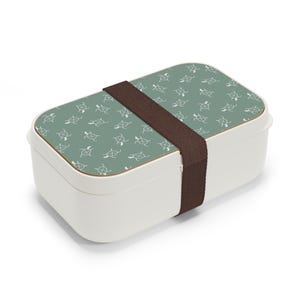 May include: A white lunchbox with a green lid featuring a repeating pattern of white turtle silhouettes. The lunchbox has a brown strap.
