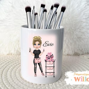 May include: White ceramic makeup brush holder featuring a cartoon illustration of a woman named Evie. The holder is filled with silver makeup brushes. The illustration includes a makeup cart and the text "Evie".
