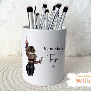 May include: White ceramic makeup brush holder with a black and white illustration of a woman holding makeup. The text reads "Beautician Tanya" with a heart symbol. The holder contains several makeup brushes.