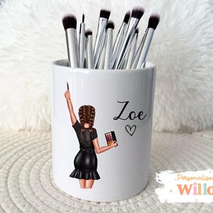 May include: White ceramic makeup brush holder with a cartoon illustration of a person holding a makeup brush and palette. The name "Zoe" and a heart are printed on the holder. The holder is filled with makeup brushes.