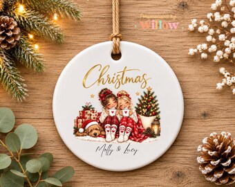 May include: A round white ceramic Christmas ornament hanging from a tan rope. The ornament features a gold script text reading Christmas, an illustration of two people with a dog, wrapped gifts, a tree, and the custom name text Molly & Lucy.