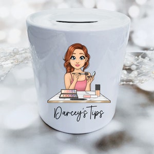 May include: White ceramic money box featuring a cartoon illustration of a woman applying makeup. The illustration includes makeup brushes, eyeshadow palettes, and foundation. The text "Darcey's Tips" is printed on the front of the money box.