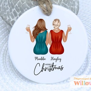 May include: A white ceramic Christmas bauble with two figures in teal and red dresses, holding wine glasses. The names "Maddie" and "Hayley" are displayed, with "Merry Christmas" written below. A jute string is attached for hanging.