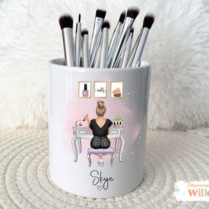 May include: White ceramic makeup brush holder with a decorative illustration of a person at a vanity. The holder contains several makeup brushes. The design includes the name "Skye" and the word "nails" on the holder.