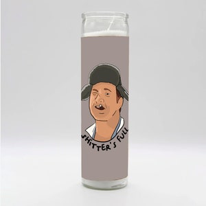 May include: A white candle with a gray label featuring a cartoon illustration of a man wearing a winter hat and a mustache. The text on the label reads "Shitter's Full".