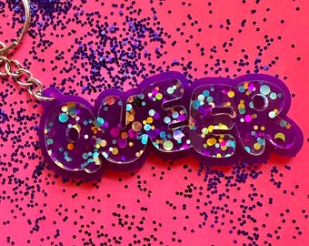 May include: A purple keychain with the word "QUEER" in bubble letters, filled with colorful glitter. The keychain is on a silver chain and sits on a pink surface scattered with purple glitter.
