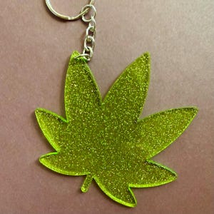 May include: A glittery green cannabis leaf keychain. The keychain features a five-pointed leaf shape with a silver-colored chain and keyring. The leaf has a sparkly, textured appearance.