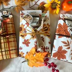 May include: Three gift-wrapped reading log sets with autumn patterns. One is plaid, another has sunflowers, and the third has fall leaves. Each is tied with a ribbon and includes a bookmark, pen, and a Galaxy chocolate bar.