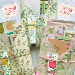 May include: Self-care gift sets wrapped in floral paper. Each set contains items like hand cream tubes, notebooks, and tea. The sets are tied with twine and feature the text "Self Care" in a heart-shaped bubble.