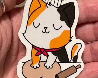 May include: A calico cat wearing a red bandana and a chef's hat is kneading dough with a rolling pin. The cat has its eyes closed and a happy expression on its face.