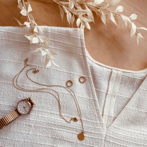 May include: A white linen top with a V-neckline and a gold chain necklace with a heart charm and a coin pendant. The necklace is layered over a gold chain bracelet with a watch.