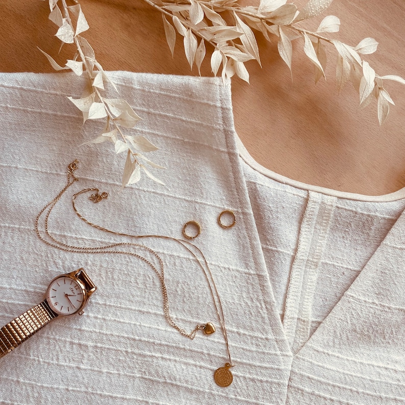 May include: A white linen top with a V-neckline and a gold chain necklace with a heart charm and a coin pendant. The necklace is layered over a gold chain bracelet with a watch.