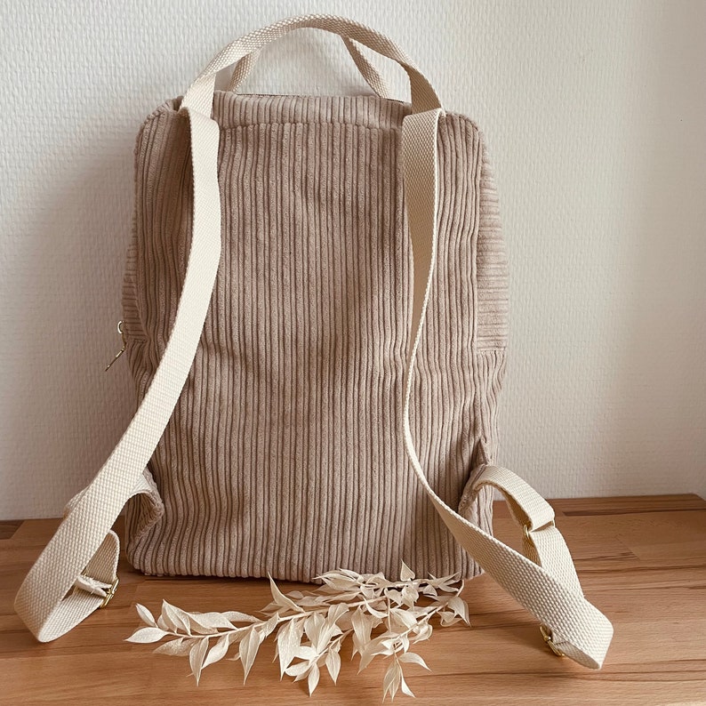 K&ouml;nnte beinhalten: Ein beigefarbener Cordrucksack mit wei&szlig;en Tr&auml;gern. Der Rucksack hat einen Rei&szlig;verschluss an der Vorderseite und ist aus einem weichen, strukturierten Stoff gefertigt.