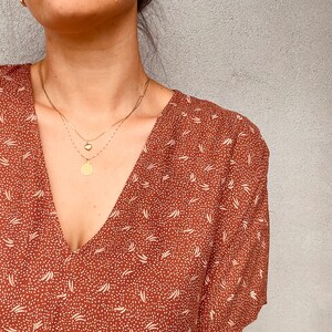 May include: A woman wearing a brown and white patterned dress with a gold necklace featuring a heart charm and a round pendant.