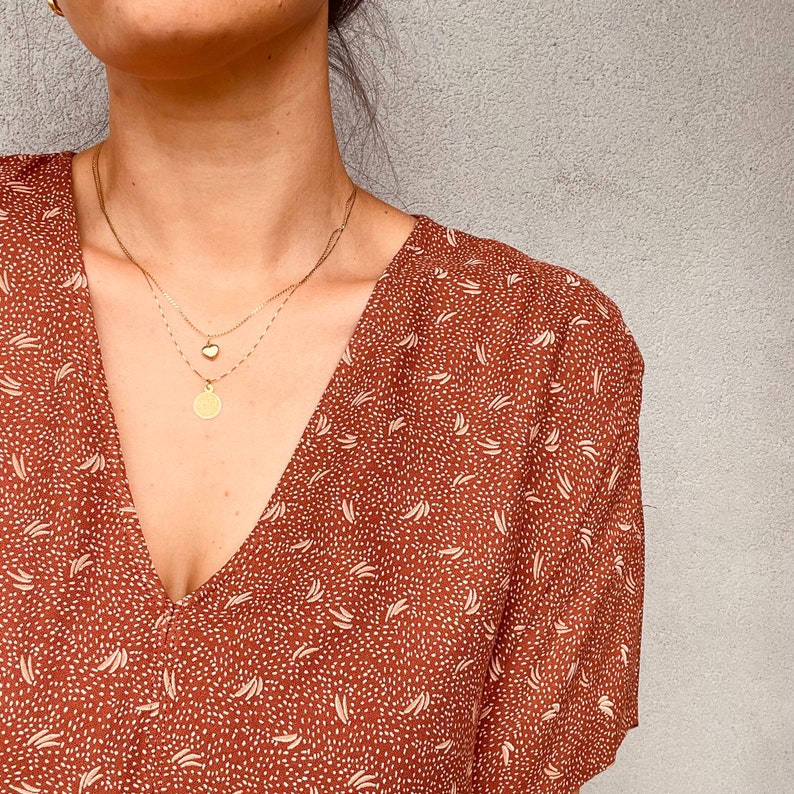 May include: A woman wearing a brown and white patterned dress with a gold necklace featuring a heart charm and a round pendant.