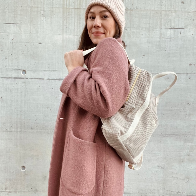 K&ouml;nnte beinhalten: Eine Frau tr&auml;gt einen rosa flauschigen Mantel und eine wei&szlig;e Strickm&uuml;tze und tr&auml;gt einen cremefarbenen Cordrucksack mit wei&szlig;en Tr&auml;gern.