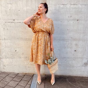 May include: A woman wearing a gold and white floral print midi dress with a gathered waist and short sleeves. She is holding a straw tote bag with a sprig of greenery.