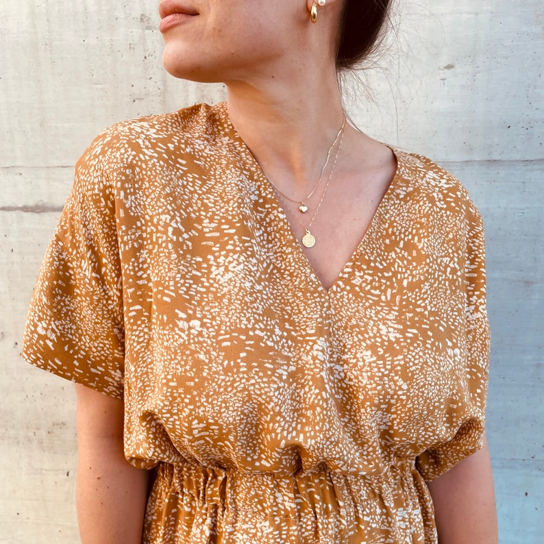 May include: A mustard yellow dress with a white abstract print. The dress has a V-neckline and short sleeves. The model is wearing a gold necklace with a small heart pendant and a larger round pendant.