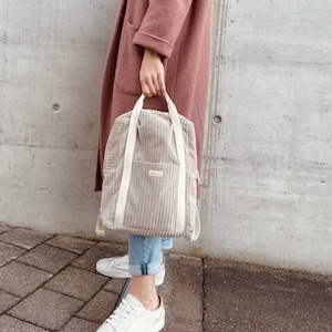 K&ouml;nnte beinhalten: Ein beigefarbener Cordrucksack mit Kordelzugverschluss und zwei verstellbaren Tr&auml;gern. Der Rucksack hat eine kleine Tasche auf der Vorderseite und einen Rei&szlig;verschluss an der Seite.