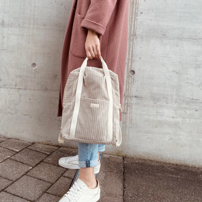 K&ouml;nnte beinhalten: Ein beigefarbener Cordrucksack mit Kordelzugverschluss und zwei verstellbaren Tr&auml;gern. Der Rucksack hat eine kleine Tasche auf der Vorderseite und einen Rei&szlig;verschluss an der Seite.
