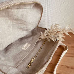 K&ouml;nnte beinhalten: Eine beige Leinen-Tragetasche mit einer Rei&szlig;verschlusstasche und einem goldenen Rei&szlig;verschluss. Die Tasche hat ein kleines Etikett mit der Aufschrift "Little Linen Co."