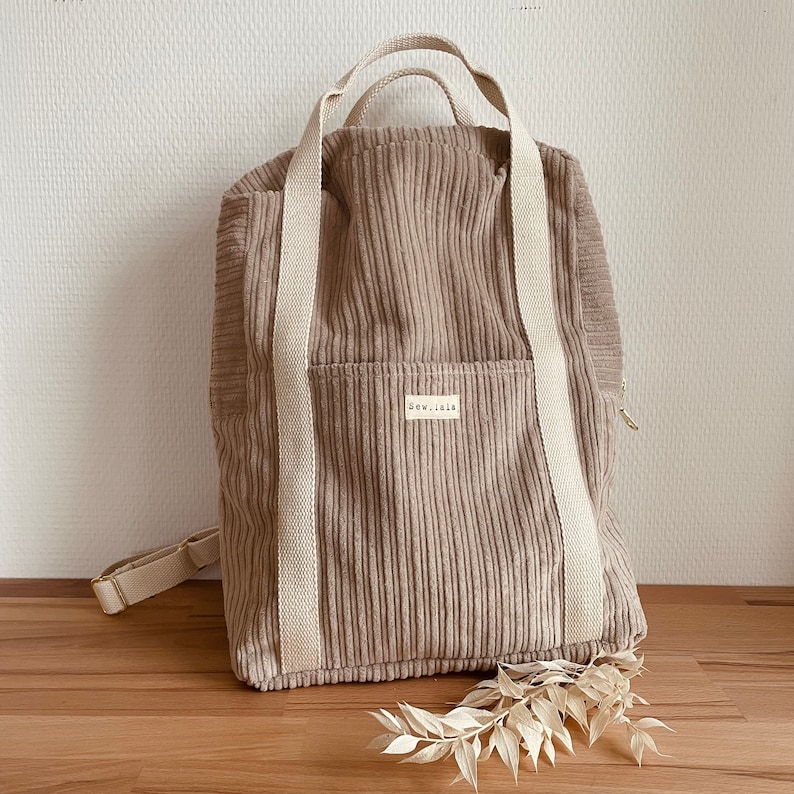 K&ouml;nnte beinhalten: Ein beigefarbener Cordrucksack mit wei&szlig;en Tr&auml;gern und einer kleinen Tasche auf der Vorderseite. Die Tasche hat ein wei&szlig;es Etikett mit dem Text "sew.isola" darauf.