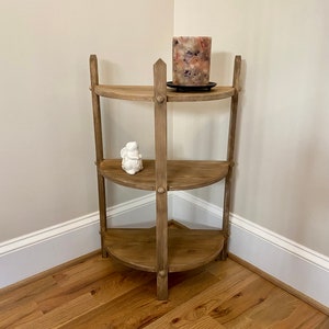 May include: A wooden corner shelf with three shelves. The shelf is made of natural wood and has a rustic finish. The top shelf has a decorative candle holder with a pink and white candle. The middle shelf has a white ceramic figurine. The bottom shelf is empty.