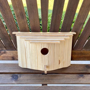 May include: A natural wood birdhouse with a round entry hole and a small perch. The birdhouse is rectangular with a curved bottom and is set against a wooden bench and a brown wooden fence.