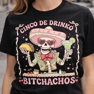 May include: Black t-shirt with a festive skeleton graphic. The skeleton wears a pink sombrero and heart-shaped sunglasses, holding a taco and a margarita. Text reads "CINCO DE DRINKO" and "BITCHACHOS" in pink.